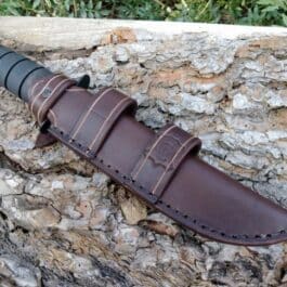 A Ka Bar Fighting Knife with a Leather Scout Sheath laying on top of a rock.