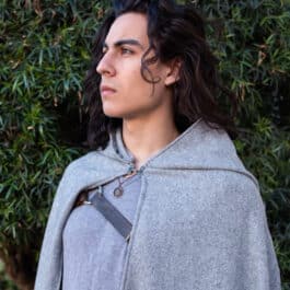 A man in a wool hooded cloak standing in front of a bush.