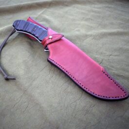 a Handmade Leather Sheath for the TOPs Longhorn Bowie with a black handle laying on a bed.
