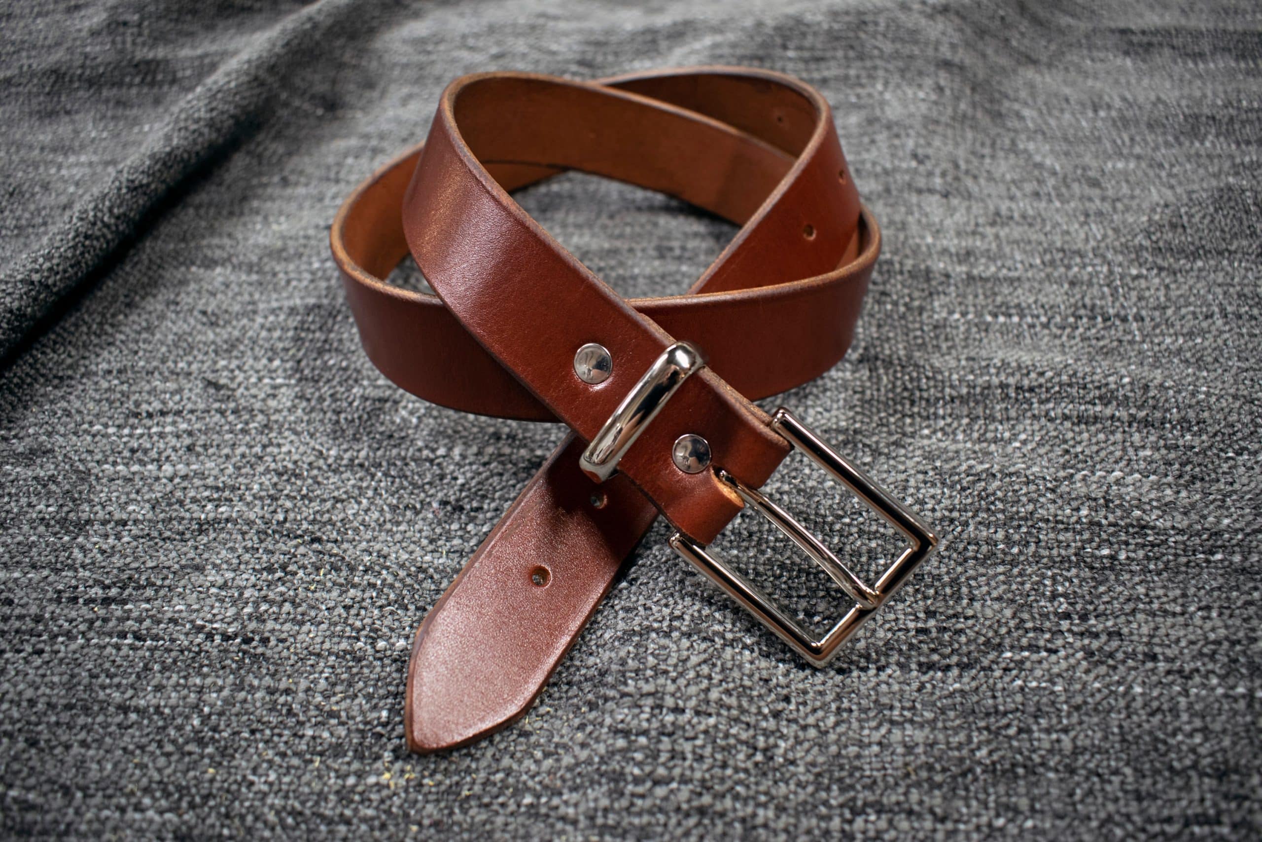 A Handmade Leather Dress Belt is laying on a gray cloth.