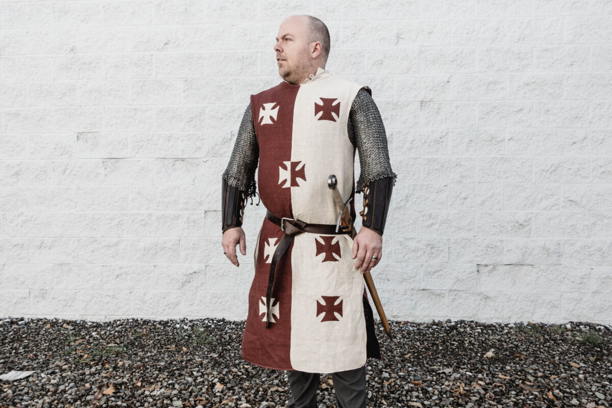 a man wearing a burgundy and cream linen surcoat with crosses on the front