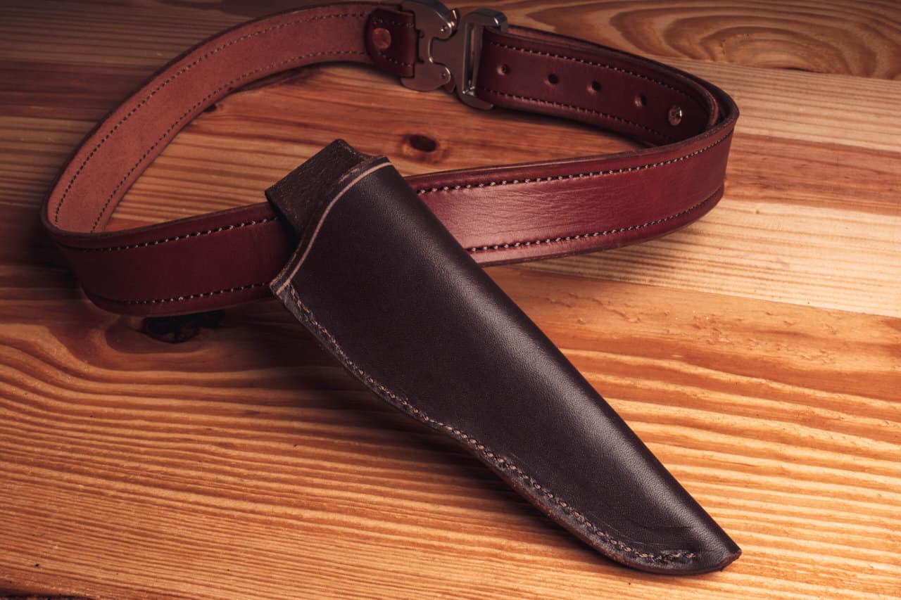 A Handmade Leather Sheath attached to a leather belt on a wooden surface.
